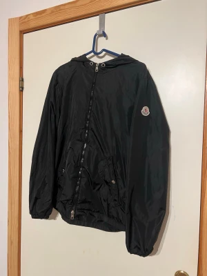 Svart vindjacka från Moncler - Snygg svart vindjacka från Moncler med huva och dragkedja framtill. Jackan har två fickor med knappstängning och Moncler-logga på ärmen. Materialet är tunt och glansigt, perfekt för blåsiga dagar. Huvan har en röd och vit rand som detalj. Perfekt för den som vill klä upp sig till ett bra pris!! Hör av er vid funderingar!!