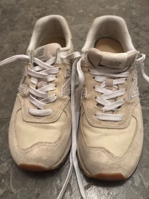 Beige New Balance sneakers - Snygga beige och vita sneakers från New Balance med klassisk N-logga på sidan. Skorna har ovandel i mocka och textil, vita skosnören och gummisula. Perfekta för en avslappnad och trendig stil.