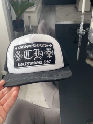 Chrome Hearts svartvit truckerkeps - Snygg svartvit keps från Chrome Heartf med stort broderat patch framtill med logga och texten 'HOLLYWOOD, USA'. Klassisk trucker-modell med mesh baktill och böjd skärm. Perfekt för dig som gillar streetwear och statement-accessoarer.