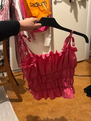 Rosa smockad linnetopp med volang - Supersöt rosa linnetopp med smockad midja och volangkant. Axelbanden är knytbara för en personlig passform och toppen har en glansig finish. Perfekt för dig som gillar färg och trendiga detaljer.