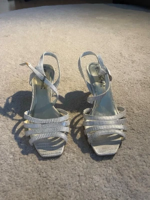 Silvriga sandaletter med remmar - Säljer ett par silvriga sandaletter från Jackie med smala remmar och öppen tå. Sandalerna har en liten klack och spänne vid ankeln. Materialet har en glansig, texturerad finish som ger en lyxig känsla. Perfekta för dig som vill ha något stilrent och trendigt.