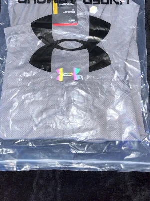 Grå Under Armour träningströja - Snygg grå träningströja från Under Armour med stort svart logotryck på bröstet och en mindre logga i regnbågsfärger. Tillverkad i ett lätt och ventilerande funktionsmaterial, perfekt för träning och sportaktiviteter.