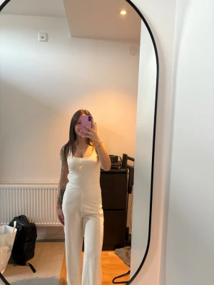 Vit jumpsuit - Säljer en vit jumpsuit med smala axelband och raka, vida ben. Ganska tjockt tyg vilket gör att den inte är så genomskinlig. Har en dragkedja längst sidan