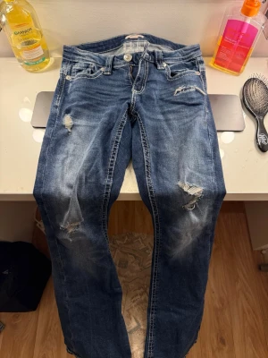 Blå slitna jeans med broderade fickor - Säljer ett par blå jeans med slitningar och hål på benen för en trendig look. Jeansen har raka ben och snygga broderade detaljer på backfickorna.