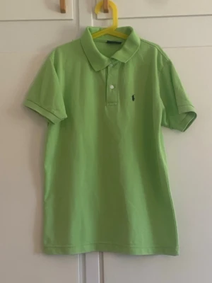 Grön pikétröja från Ralph Lauren - Snygg ljusgrön pikétröja från Ralph Lauren med klassisk krage och två knappar framtill. Tröjan har korta ärmar och den ikoniska lilla loggan broderad på bröstet. Perfekt till sommaren för en fräsch och sportig look.