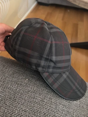 Mörkgrå rutig keps från Burberry - Säljer en snygg mörkgrå keps från Burberry med klassiskt rutmönster i svart, grått och röda linjer. Kepsen har böjd skärm och justerbar rem bak. Perfekt accessoar för en trendig streetwear-look.