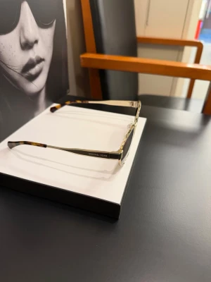 Guldiga solglasögon Michael Kors - Snygga solglasögon från Michael Kors med smal, rektangulär form och guldfärgade metallbågar. Glasen är lätt tonade i brunt och skalmarna har en diskret sköldpaddsmönstrad detalj. Perfekt accessoar för dig som vill ha en stilren och trendig look.