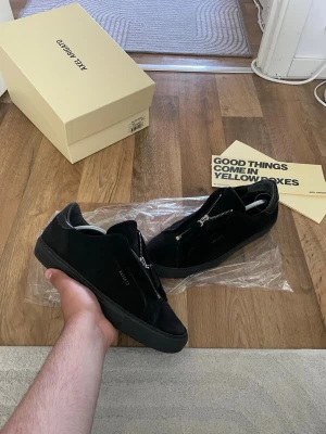 Svarta sneakers från Axel Arigato zip 90 - Snygga svarta sneakers från Axel Arigato i mocka med coola silverfärgade dragkedjor istället för vanliga snören. Skorna har en chunky svart sula och minimalistisk design med diskret logga på sidan. Perfekta för dig som gillar stilrena och moderna detaljer.