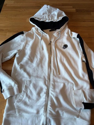 Vit hoodie med svarta detaljer från La Vichy - Snygg vit hoodie med dragkedja från La Vichy. Har svarta detaljer längs ärmarna och svart insida på huvan. Utrustad med två stora fickor fram och justerbar dragsko i huvan. Liten broderad logga på bröstet. Har nästan aldrig använt för den inte passat mig. Org pris 1700kr