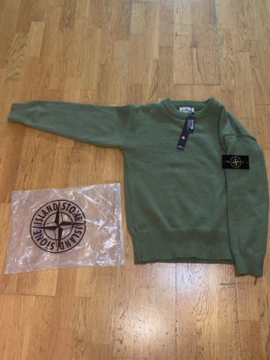 Grön stickad tröja från Stone Island - Snygg grön stickad tröja från Stone Island med rund halsringning och ribbade muddar. Tröjan har en klassisk patch med knappdetalj på ärmen och är tillverkad i ett mjukt, värmande material. Perfekt för dig som gillar streetwear och stilren design.