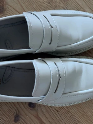 Vita loafers i skinn - Säljer ett par stilrena vita loafers i slätt skinn med klassisk penny strap och rundad tå. Skorna har en diskret sula och minimalistisk design som passar till många olika outfits. Perfekta för dig som gillar en clean och modern look.