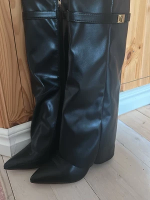 Svarta knähöga boots - Säljer ett par svarta knähöga stövlar i glansigt skinnimitation med spetsig tå och bred klack. Stövlarna har ett dekorativt spänne upptill och dragkedja på insidan. Perfekta för dig som vill ha en edgy och trendig look. Säljer eftersom de var för små.