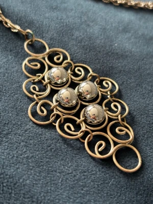 Vintage Halsband - Unikt halsband i vintage-stil med en stor, dekorativ hängare i metall. Hänget har fyra blanka metallpärlor omgivna av snirkliga detaljer. Kedjan är tunn och matchar hängets färg. Perfekt för dig som gillar retro och statement-smycken. I fint skick. Ingen stämpel. Minimal oxidering på vissa ställen.  Ungefär 39 cm långt.