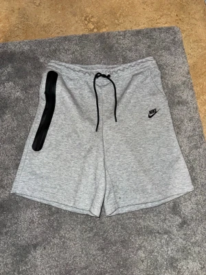 Nike tech shorts grå - Snygga grå shorts från Nike med svart snörning i midjan och en svart dragkedjedetalj på vänster sida. Tillverkade i mjuk bomullsmix och har en avslappnad passform. Perfekta för sommaren eller träning.