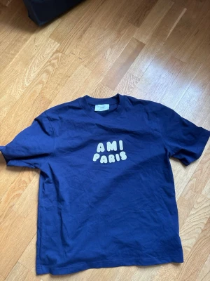 Blå t-shirt från Ami Paris - Snygg mörkblå t-shirt från Ami Paris med broderad text 'AMI PARIS' i vitt framtill. Klassisk passform och rund halsringning. Tillverkad i mjuk bomull som känns skön mot huden. Perfekt för en avslappnad och trendig look.