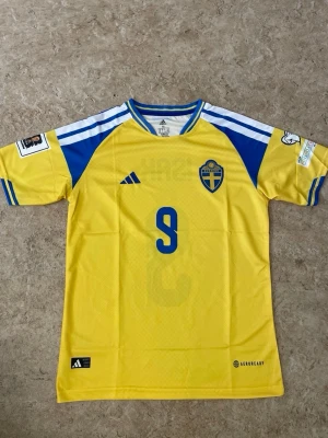 Sverige matchtröja Isak #9 Adidas - Gul och blå Sverige matchtröja från Adidas med Isak och nummer 9 på ryggen. Tröjan har officiella landslagsmärket på bröstet, blå detaljer på axlar och ärmar samt Aeroready-material som andas. Perfekt för fotbollsfans!