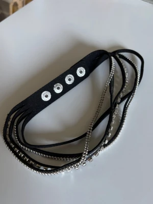 Svart halsband med silverdetaljer - Snyggt svart halsband med flera tunna band i mockaimitation och silverfärgade kedjor samt små pärlor. Halsbandet har tryckknappsstängning med fyra silverfärgade knappar för justerbar storlek. Perfekt accessoar för en trendig look.
