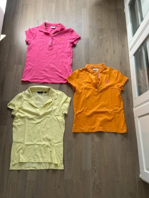Tre pikétröjor i olika färger - Säljer tre pikétröjor i bomull: en ljusgul från Gant, en orange och en rosa från Ralph Lauren. Alla har klassisk krage, knappar framtill och broderad logga på bröstet. Perfekta till sommaren och ger en fräsch look till din outfit.