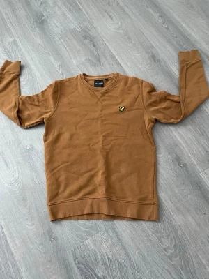 Brun sweatshirt från Lyle & Scott - Snygg brun sweatshirt från Lyle & Scott med klassisk rund halsringning och gul logga på bröstet. Tröjan har ribbade muddar vid ärmslut och nederkant. Perfekt för en avslappnad och stilren look.