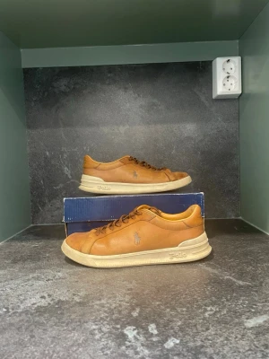Bruna Polo Ralph Lauren sneakers - Snygga bruna sneakers från Polo Ralph Lauren i läder med klassisk låg profil och vita sulor. Ikoniska Polo-loggan på sidan och diskreta sömmar ger en clean look. Perfekta för dig som gillar stilrena och tidlösa skor.