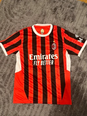 AC Milan matchtröja röd/svart - Säljer en AC Milan matchtröja i rött och svart med vita detaljer. Tröjan har klubbmärke på bröstet, sponsortryck 'Emirates Fly Better' och klassiska vertikala ränder. Tillverkad i lätt och ventilerande syntetmaterial, perfekt för fotbollsträning eller match.