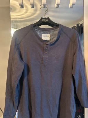 Blågrå långärmad henley från Braintree - Snygg långärmad henley-tröja från Braintree med tre knappar vid halsen. Tröjan har en avslappnad passform och är tillverkad i ett mjukt, tunt tyg som känns skönt mot huden. Perfekt för en chill och stilren look.