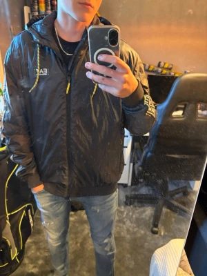 Svart EA7 vindjacka med huva Armani. - Snygg svart vindjacka från EA7 Emporio Armani med huva och gula detaljer på snörena. Jackan har logga på bröstet och ärmen samt dragkedja framtill. Tillverkad i glansigt, lätt material som passar perfekt till streetwear-stilen. Jackan är använd max 2 ggr. Nypris 2600