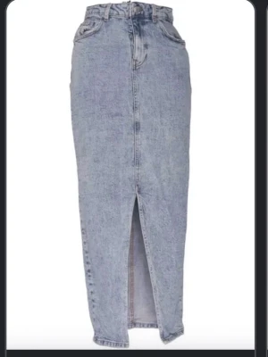 Lång jeanskjol med slits framtill - Säljer en klassisk lång jeanskjol i blå denim med hög midja och slits framtill. Kjolen har femficksmodell, bälteshällor och stängs med dragkedja och knapp. Perfekt till sneakers eller boots för en cool vardagslook.