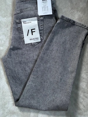 Grå slim tapered jeans Selected Femme - Snygga grå jeans från Selected Femme i slim tapered fit. Jeansen har klassisk femficksdesign och är tillverkade av ekologisk bomull. Perfekta för dig som gillar en smalare passform med avsmalnande ben.