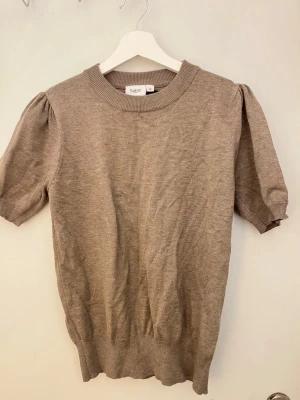 Beige stickad topp från Saint - Stickad topp i beige från Saint med puffärm och ribbade muddar. Kort ärm och rund halsringning ger en clean look. Perfekt till jeans eller kjol för en enkel men snygg outfit. Materialet känns mjukt och lätt, perfekt för lager på lager. Storlek M men passar S