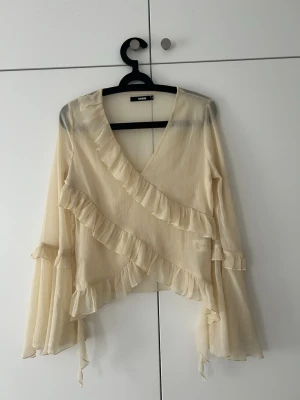 Krämvit volangblus från BikBok - Säljer en krämvit blus från BikBok med omlottmodell och volangdetaljer längs ringningen och framtill. Blusen har långa, vida ärmar och är gjord i ett tunt, lätt och något transparent tyg som ger ett flowy intryck. Perfekt för dig som gillar romantisk och bohemisk stil.