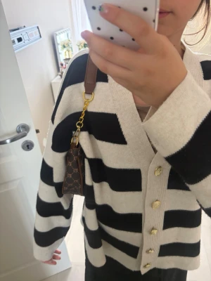 Brun handväska med monogram - Snygg brun handväska från mixxini & bags med klassiskt monogrammönster och guldfärgade detaljer. Väskan har dragkedja upptill och en axelrem i brunt läder. Perfekt accessoar för att lyfta din outfit.