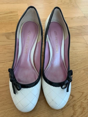 Vita pumps med svarta detaljer Love Like Lip - Säljer ett par eleganta pumps i storlek 36, i cremevitt med svart lackkant och dekorativ rosett framtill. Klassisk och feminin modell som passar både till fest och vardag.  I bra använt skick. Endast använda inomhus. Bekväm passform. Klackhöjd ca 6–7 cm.