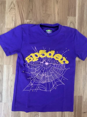 Lila Sp5der t-shirt med spindelnätsprint - Cool lila t-shirt med ett stort spindelnätsmotiv och texten 'sp5der' i gul, bubblig stil över bröstet. T-shirten har korta ärmar och är tillverkad i mjuk bomull. Perfekt för dig som gillar unika och färgstarka plagg.