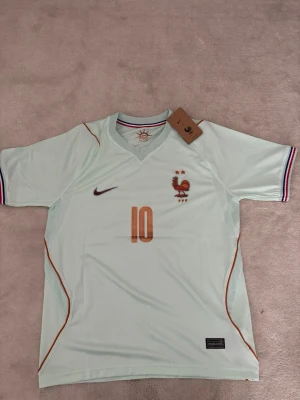 Frankrike Nike fotbollströja #10 M och S - Ljusblå fotbollströja från Nike med Frankrikes landslagsemblem och nummer 10 i orange framtill. Tröjan har korta ärmar med ränder i blått, vitt och rött vid ärmsluten samt broderad tupp och två stjärnor på bröstet. Tillverkad i lätt och ventilerande material.