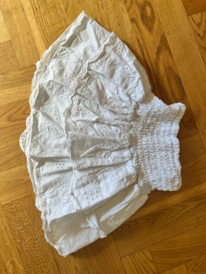Vit kort kjol med broderi från H&M - Söt vit kort kjol från H&M med broderade blommönster och bred smock i midjan. Kjolen har volangkant och är gjord i ett lätt bomullstyg som passar perfekt till varma dagar.