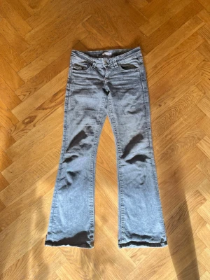 Grå bootcut jeans från Ginatricot - Säljer ett par grå bootcut jeans från Ginatricot med klassisk femficksmodell och knappgylf. Jeansen har en snygg tvättad look och utsvängda ben som ger en retrovibe. Perfekta för dig som gillar en avslappnad men trendig stil.
