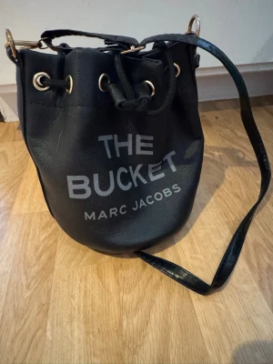 Svart bucket bag Marc Jacobs - Snygg svart axelremsväska från Marc Jacobs i bucket-modell. Väskan har dragskostängning med guldfärgade detaljer och texten 'THE BUCKET MARC JACOBS' på framsidan. Tillverkad i strukturerat konstläder och har justerbar axelrem.