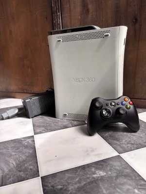 Xbox 360 spelkonsol med handkontroll - Säljer en klassisk Xbox 360 spelkonsol i vit plast med tillhörande svart trådlös handkontroll och strömadapter. Konsolen har en stilren, rundad design och är perfekt för gamingkvällar med vänner. Passar dig som gillar retrospel och vill ha en ikonisk konsol i samlingen.