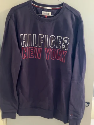 Mörkblå Hilfiger sweatshirt - Mörkblå sweatshirt från Tommy Hilfiger med trycket 'HILFIGER NEW YORK' i vitt och rött på bröstet. Klassisk rund halsringning och långa ärmar. Perfekt för en avslappnad och trendig stil.