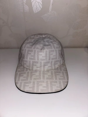 Fendi keps - vit - Säljer en vit keps från Fendi med det klassiska FF-monogrammet över hela kepsen. Kepsen har böjd skärm och är tillverkad i ett tygmaterial med diskret mönster. Perfekt för dig som gillar exklusiva accessoarer och vill sticka ut.