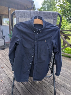 Mörkblå skjorta från Ralph Lauren stl 14-16 L - Klassisk mörkblå skjorta från Ralph Lauren med button-down krage och broderad logga på bröstet. Skjortan har långa ärmar och knappar framtill. Perfekt till jeans eller chinos för en clean och stilren look.