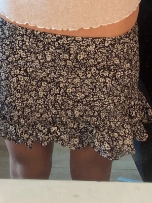 Svart blommig volangkjol - Kort svart kjol med små vita blommor och volangkant. Kjolen har en söt och ungdomlig stil, perfekt för varma dagar. Tillverkad i ett lätt och mjukt material som känns bekvämt mot huden.