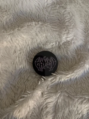 Svart popsocket med lila text - Säljer en svart popsocket till mobilen med ett coolt lila tryck i metallisk stil. Perfekt för att få bättre grepp om mobilen eller använda som ställ. Passar de flesta mobilskal och är enkel att fästa och ta bort.