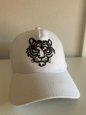 Vit keps med tigermotiv - Snygg vit keps med broderad tiger på framsidan. Tigern har svarta konturer och röda ögon vilket ger ett coolt och unikt uttryck. Kepsen har böjd skärm och klassisk passform. Perfekt accessoar för att lyfta din stil.