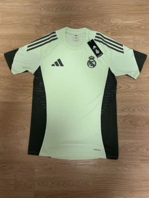 Real Madrid träningströja Adidas - Snygg ljusgrön och svart träningströja från Adidas med Real Madrid-logga på bröstet. Tröjan har korta ärmar, rund hals och klassiska Adidas-ränder på axlarna. Tillverkad i lätt och ventilerande funktionsmaterial.