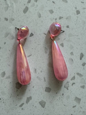 Rosa droppformade statementörhängen - Säljer ett par glansiga, droppformade örhängen i transparent rosa plast. Örhängena har en rund pärlliknande topp och en lång, avsmalnande droppe som hänger ned. Perfekta för dig som vill sticka ut med färg och form.