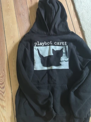 Playboi Carti svart hoodie kattmotiv - Svart hoodie med stor huva och magficka från Playboi Carti. På bröstet finns ett tryck med en svart katt och texten 'playboi carti' i vitt. Hoodien har en avslappnad passform och är gjord i mjukt bomullsmaterial. Perfekt för dig som gillar streetwear och snygga prints.