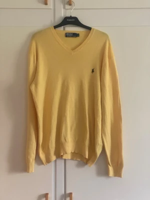 Gul v-ringad tröja från Polo Ralph Lauren - Gul v-ringad tröja från Polo Ralph Lauren med klassisk broderad logga på bröstet. Långärmad modell i mjukt material, perfekt för lager-på-lager. Enkel och stilren design som passar till många outfits.