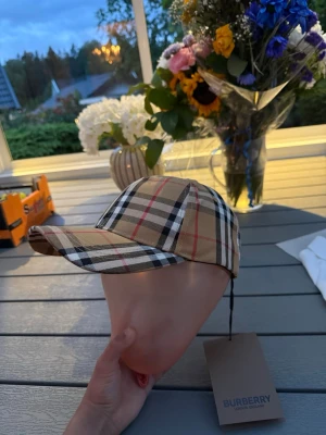 Burberry keps med klassiskt rutmönster - Snygg keps från Burberry i beige med det ikoniska rutmönstret i svart, vitt och rött. Kepsen har böjd skärm och justerbar rem baktill. Perfekt accessoar för att lyfta din stil med en lyxig touch.